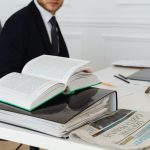 Advogado sentado à secretária, rodeado de livros jurídicos, documentos e um jornal.