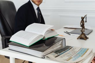 Advogado sentado à secretária, rodeado de livros jurídicos, documentos e um jornal.